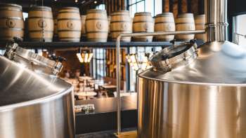 View of a modern brewery with gleaming stainless steel tanks and stacked wooden barrels in the background.