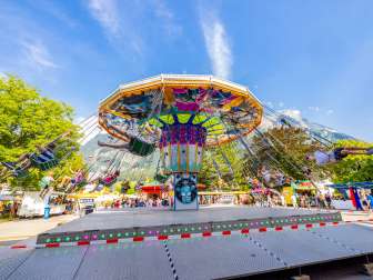Large colorful chain carousel at the fair in Balzers, full of happy visitors in front of a mountain backdrop under a blue sky.