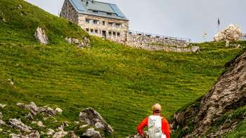 Hiker with red backpack looks at the Pfälzerhütte hut green mountain landscape.