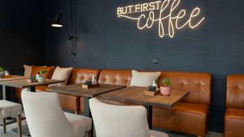 A modern café with leather sofas, white armchairs and wooden tables. The lettering "BUT FIRST coffee" shines on the dark blue wall in bright neon lettering.