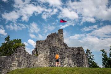 Hiker in front of the historic ruins of Schellenberg Castle with a waving Liechtenstein flag under a blue sky