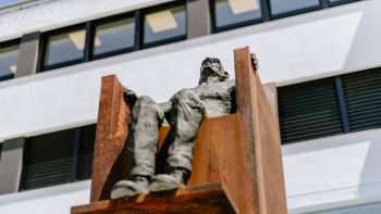 Sculpture "Hochsitz" by Robert Indermaur: Figure sits elevated on a rust-colored steel chair in front of a modern façade.