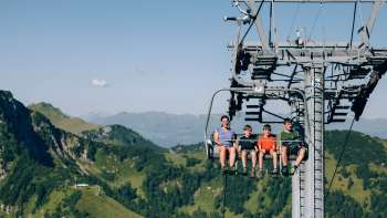 A family rides the Sareis chairlift over a green mountain landscape A family rides the Sareis chairlift over a green mountain landscape
