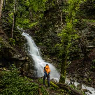 Hikers with rucksacks in front of an idyllic waterfall near Nendeln, embedded in dense forest and moss-covered rocks.