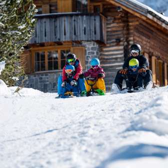 Two adults and three children sit on sledges and start a descent together in front of a snow-covered mountain hut in Malbun