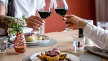 Two people toast with red wine, in the foreground a lovingly prepared main course