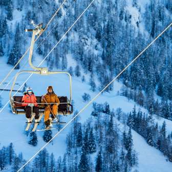 Tina Weirather and Marco Büchel sit together on a chairlift, surrounded by snow-covered trees and the winter landscape in Malbun.